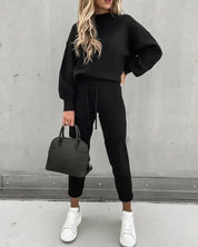 Donna in tuta nera elegante, con borsa a mano nera e sneakers bianche, posa contro sfondo grigio. Moda casual chic, abbigliamento autunnale.