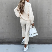 Donna in abbigliamento casual beige, con borsa bianca e scarpe da ginnastica, posa contro un muro grigio. Look moda autunnale elegante.
