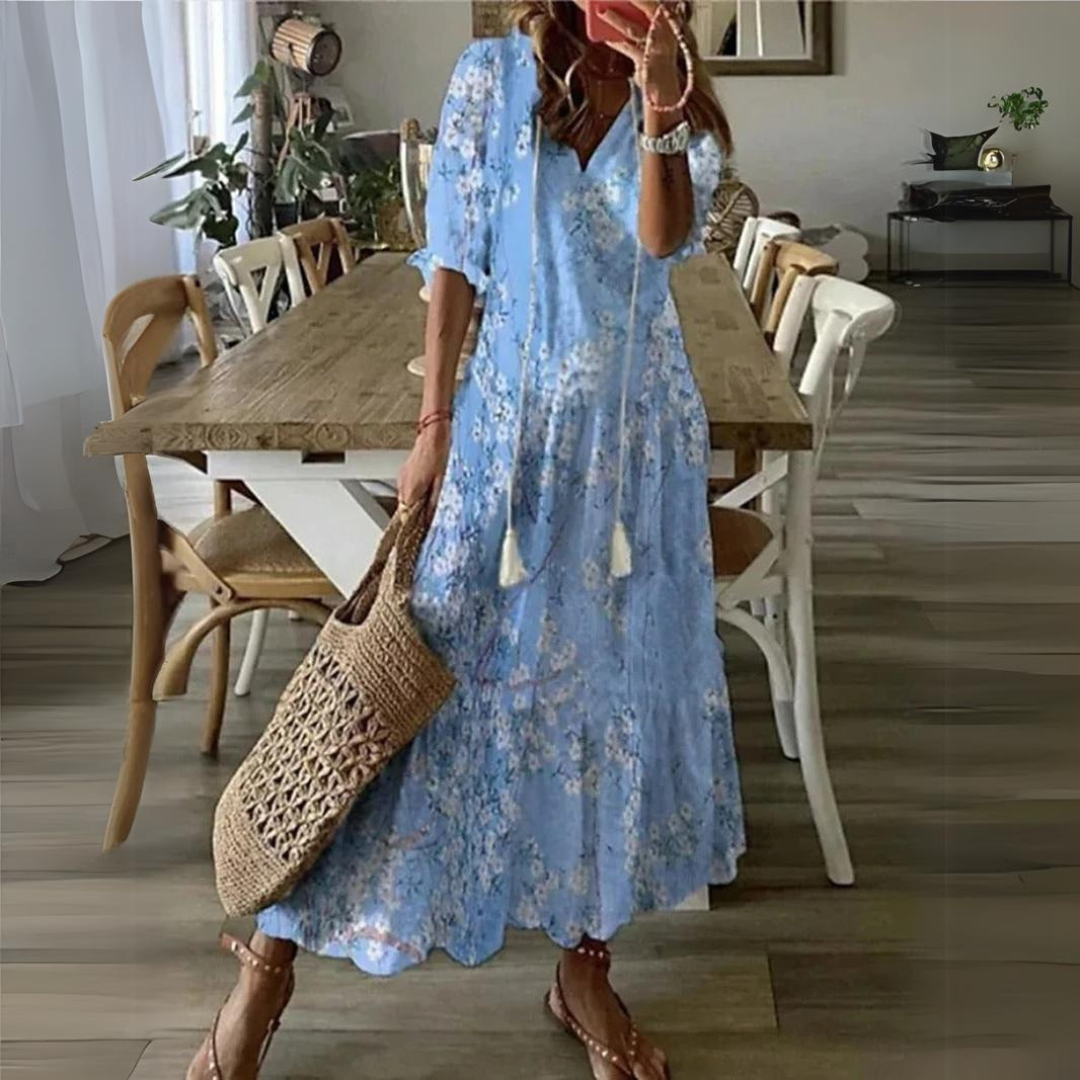 Donna con abito lungo floreale azzurro, borsa intrecciata, sandali marroni, in sala da pranzo moderna. Moda estiva, abbigliamento casual elegante.