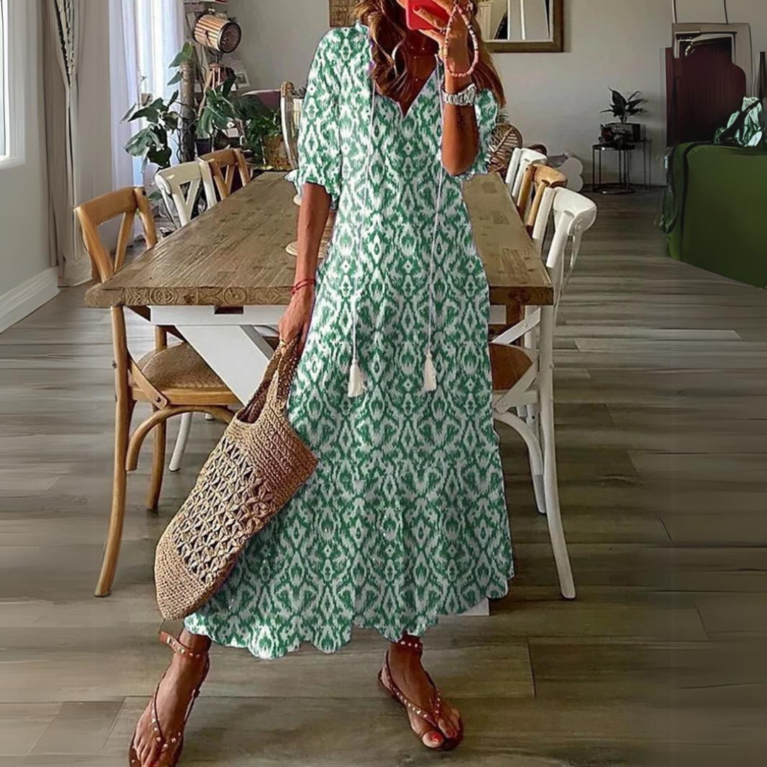Abito lungo verde con motivo ikat, maniche a tre quarti, indossato da donna con borsa intrecciata e sandali, in sala da pranzo moderna.