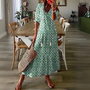 Abito lungo verde con motivo ikat, maniche a tre quarti, indossato da donna con borsa intrecciata e sandali, in sala da pranzo moderna.