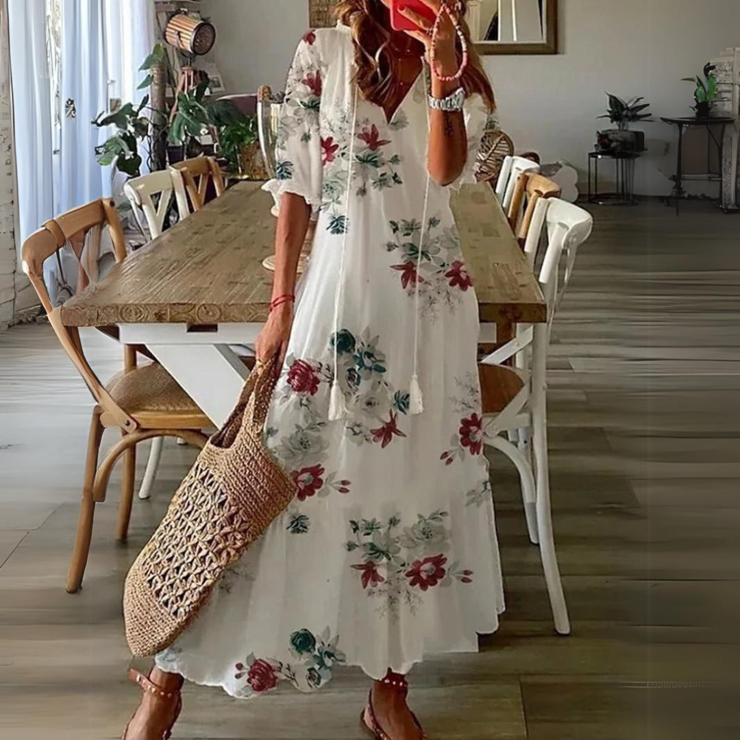 Donna con abito lungo floreale bianco, borsa di paglia, in sala da pranzo moderna. Moda estiva, abbigliamento casual elegante, arredamento interno.