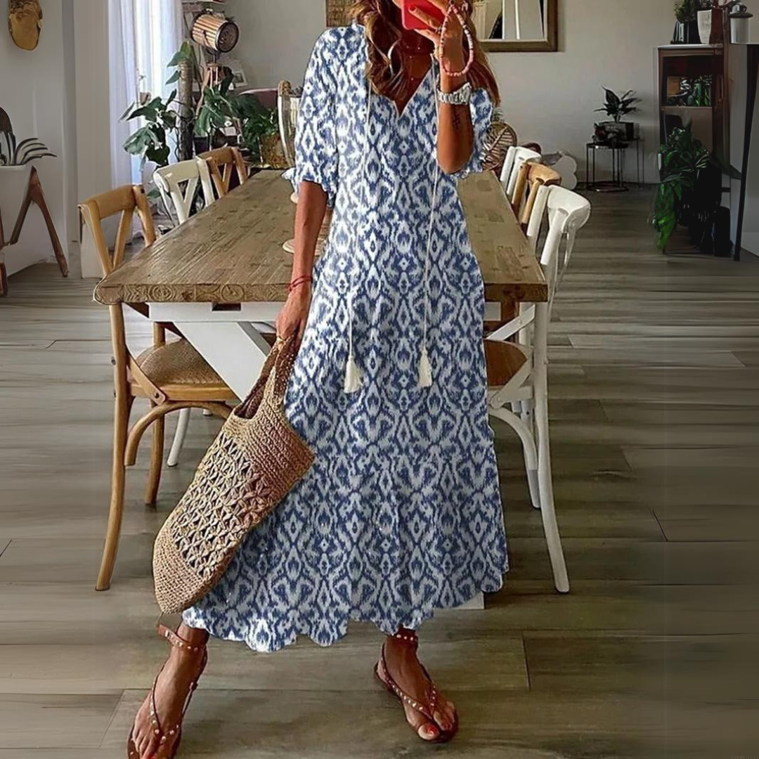 Abito lungo estivo donna con motivo azzurro e bianco, borsa in paglia, sandali marroni, ambientato in sala da pranzo moderna. Moda casual elegante.