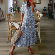 Abito lungo estivo donna con motivo azzurro e bianco, borsa in paglia, sandali marroni, ambientato in sala da pranzo moderna. Moda casual elegante.
