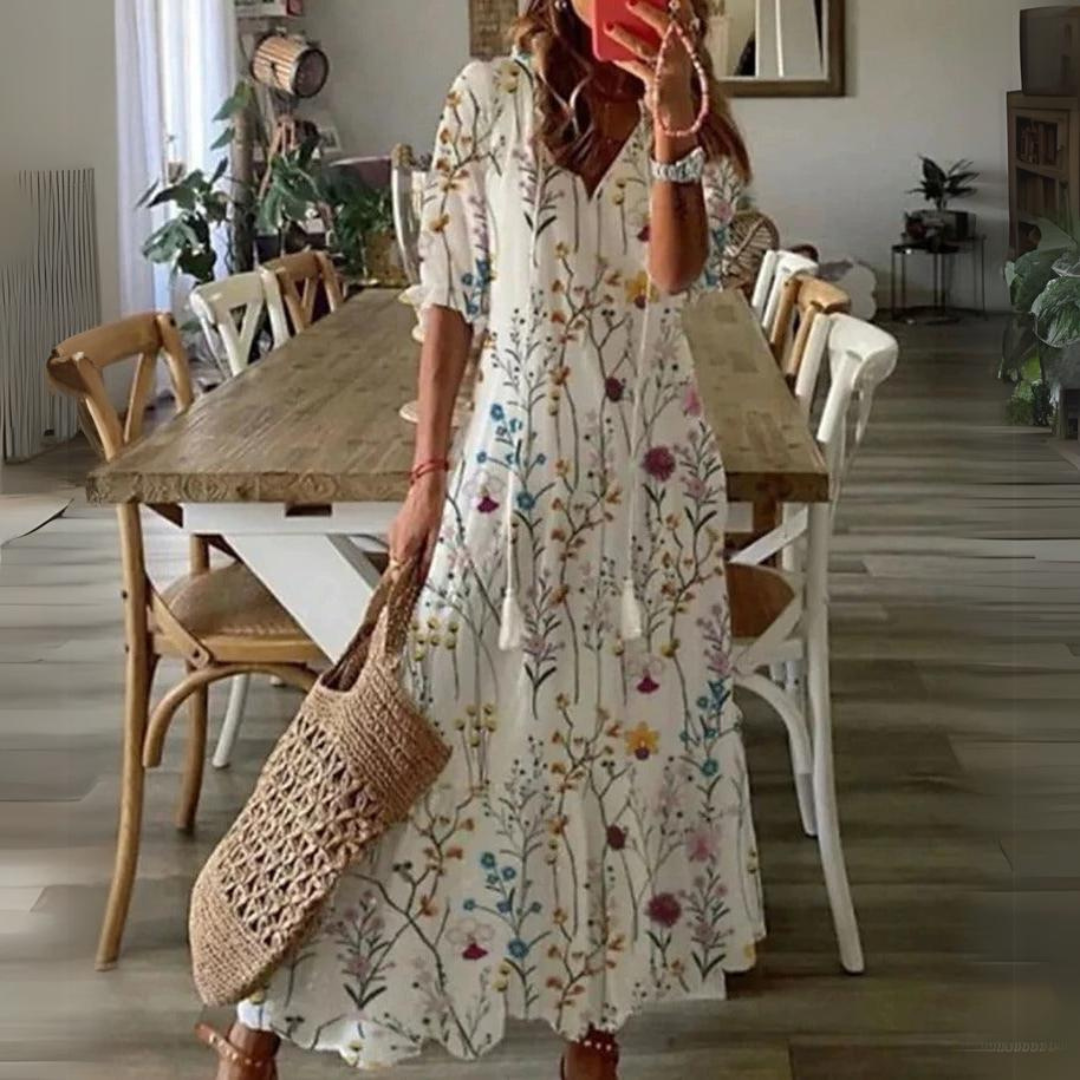 Abito lungo estivo bianco con stampa floreale colorata, maniche a tre quarti, indossato da donna con borsa in paglia, in sala da pranzo moderna.