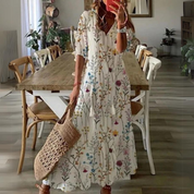 Abito lungo estivo bianco con stampa floreale colorata, maniche a tre quarti, indossato da donna con borsa in paglia, in sala da pranzo moderna.