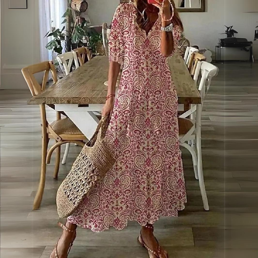 Abito lungo estivo a fantasia floreale rosa, donna con borsa di paglia, sandali, in sala da pranzo moderna con tavolo in legno e sedie bianche.