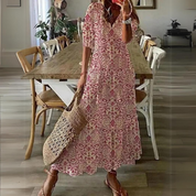 Abito lungo estivo a fantasia floreale rosa, donna con borsa di paglia, sandali, in sala da pranzo moderna con tavolo in legno e sedie bianche.