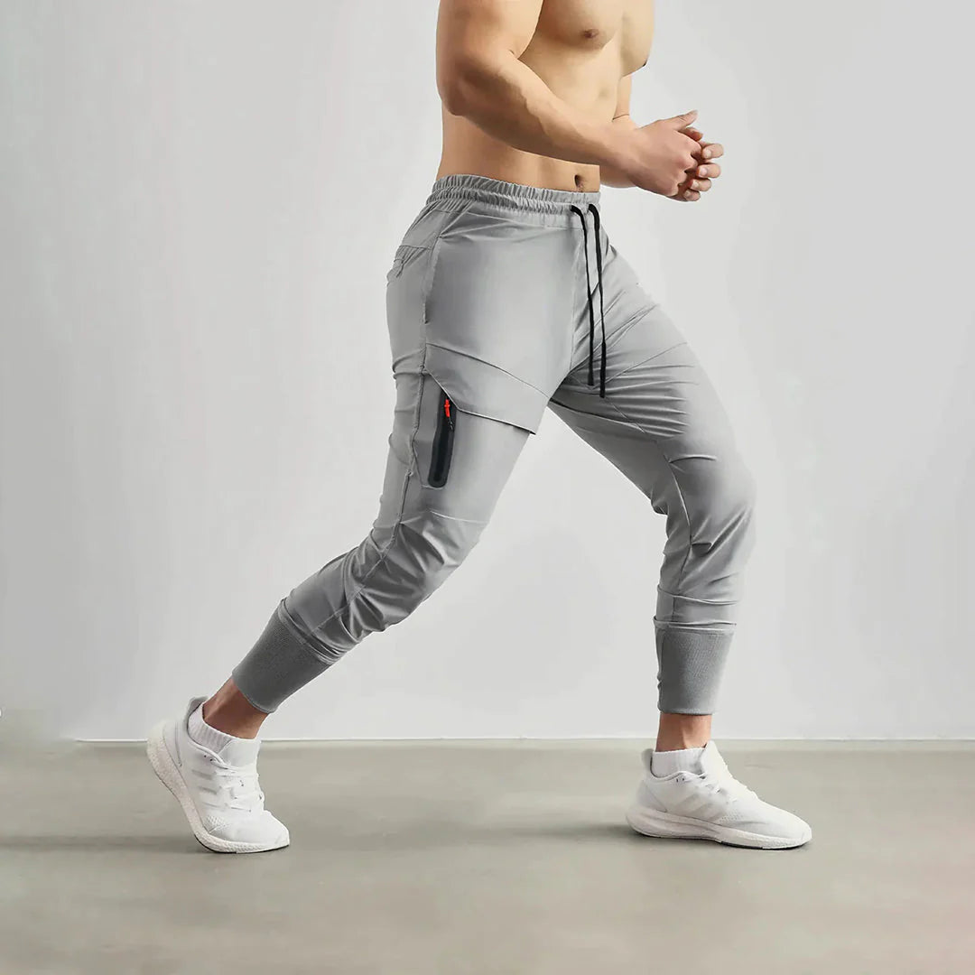 Uomo in pantaloni sportivi grigi con tasca laterale, in movimento su sfondo neutro. Abbigliamento fitness, pantaloni da jogging, moda sportiva.
