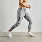 Uomo in pantaloni sportivi grigi con tasca laterale, in movimento su sfondo neutro. Abbigliamento fitness, pantaloni da jogging, moda sportiva.