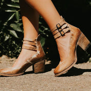 Scarpe donna in pelle marrone con tacco medio, cinturini alla caviglia e fibbie, su sfondo naturale. Eleganti e alla moda per ogni occasione.