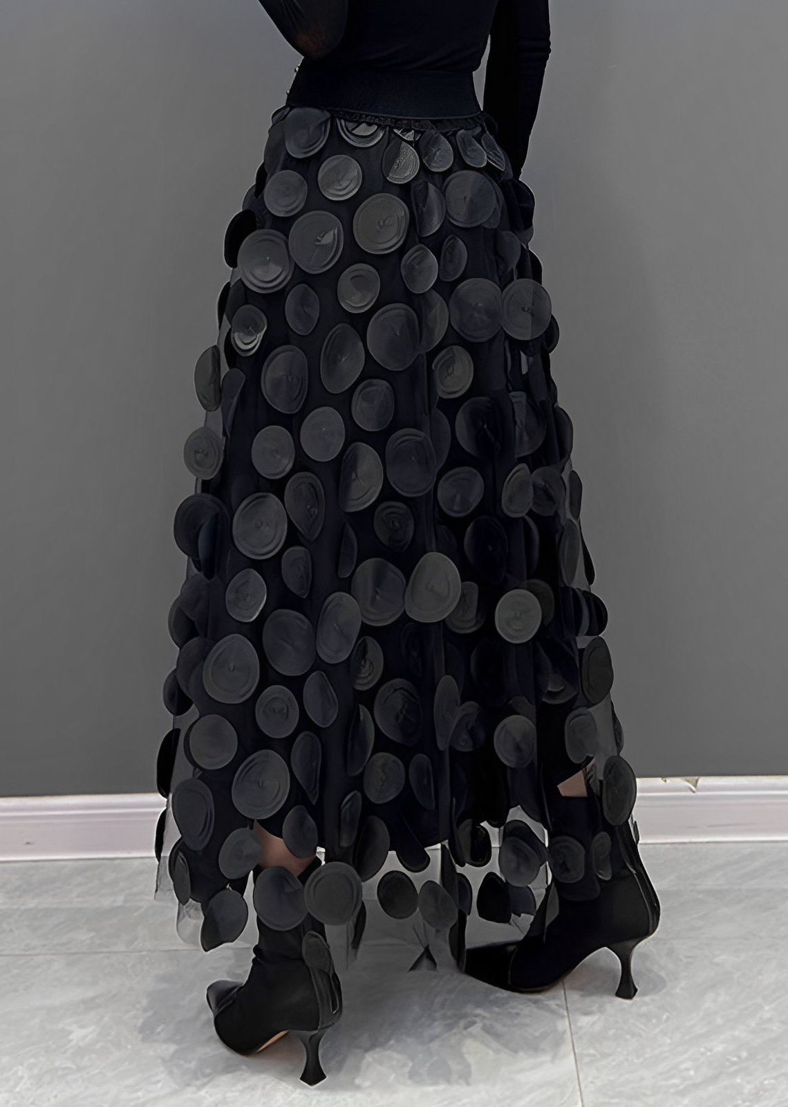 Gonna lunga nera con cerchi decorativi, design elegante e moderno, indossata con stivali neri su sfondo grigio. Moda femminile autunno inverno.