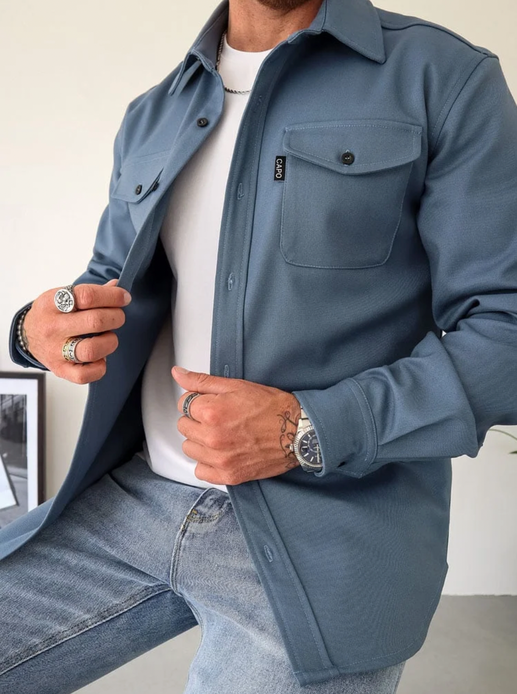Camicia uomo blu scuro, stile casual, con tasche frontali e bottoni, indossata sopra t-shirt bianca e jeans. Accessori: anelli e orologio.