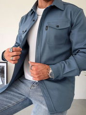 Camicia uomo blu scuro, stile casual, con tasche frontali e bottoni, indossata sopra t-shirt bianca e jeans. Accessori: anelli e orologio.