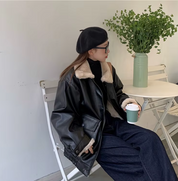 Donna con giacca in pelle nera e berretto, seduta a un tavolo bianco con pianta verde, tiene una tazza di caffè. Moda autunnale casual chic.