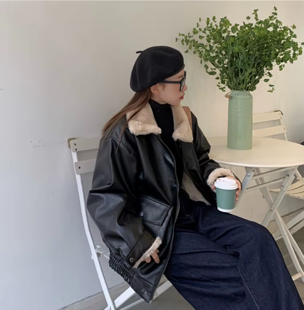 Donna con giacca in pelle nera e berretto, seduta a un tavolo bianco con pianta verde, tiene una tazza di caffè. Moda autunnale casual chic.