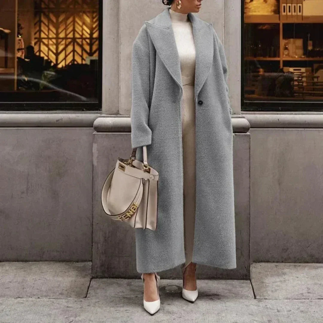 Cappotto lungo grigio elegante, donna in abito bianco, borsa beige con dettagli dorati, scarpe col tacco bianche, moda autunnale sofisticata.