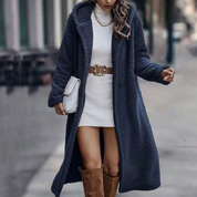 Donna con cappotto lungo blu in pelliccia sintetica, abito bianco aderente, cintura marrone, stivali alti marroni e borsa a tracolla bianca.