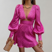 Abito corto rosa fucsia con maniche a sbuffo, scollo profondo e dettaglio cut-out, indossato con collana e borsa intrecciata. Moda elegante donna.