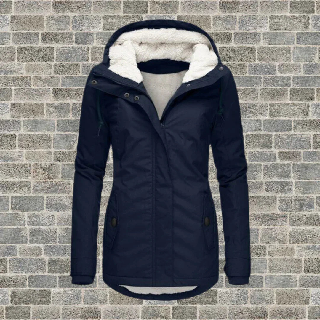 Giacca invernale donna blu con cappuccio foderato in pelliccia sintetica, chiusura a zip, tasche frontali, su sfondo muro di mattoni grigi.