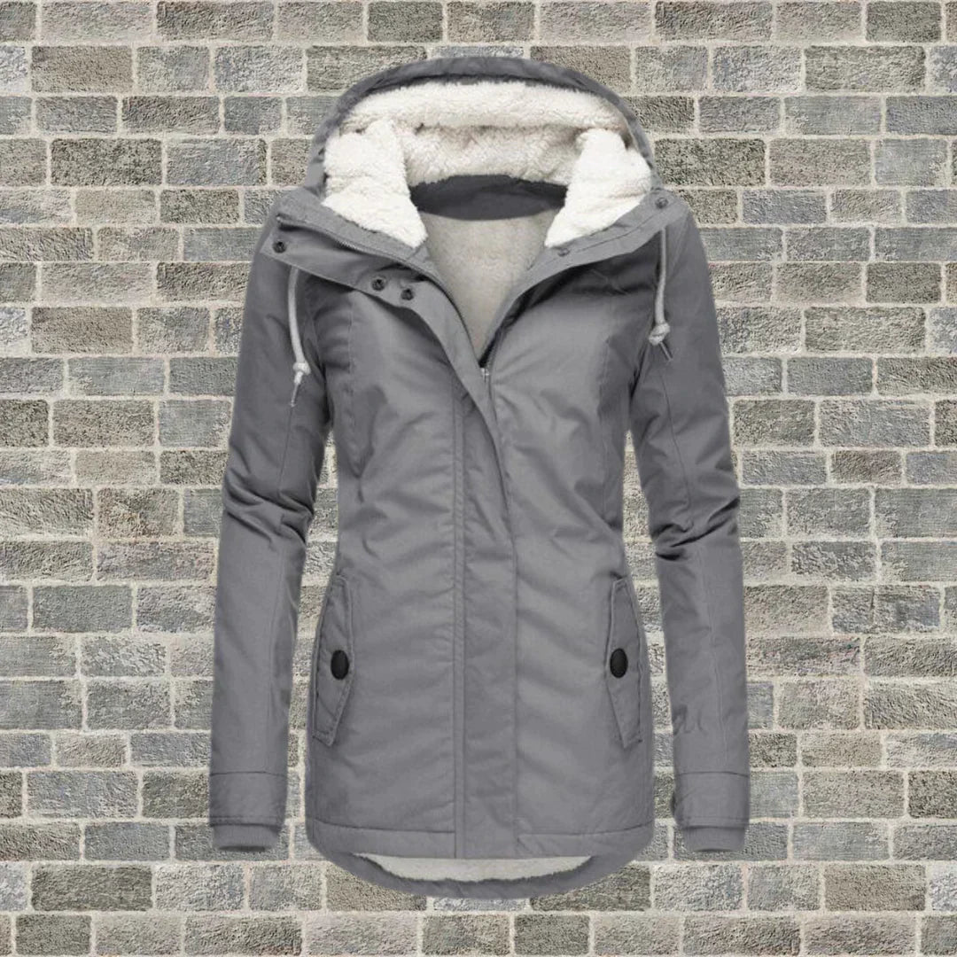 Giacca invernale grigia da donna con cappuccio foderato in pelliccia sintetica, chiusura a zip, tasche frontali, su sfondo di mattoni grigi.