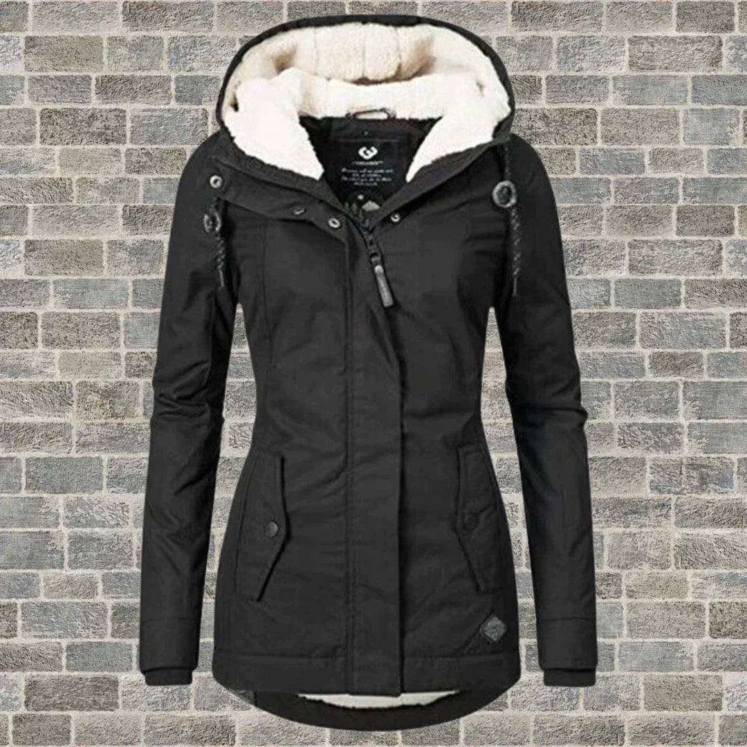 Giacca invernale nera da donna con cappuccio foderato in pelliccia sintetica, chiusura a zip, tasche frontali, su sfondo di mattoni grigi.