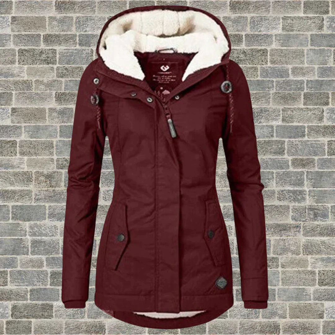 Giacca invernale da donna bordeaux con cappuccio foderato in pelliccia sintetica, chiusura a zip, bottoni decorativi, su sfondo di mattoni grigi.
