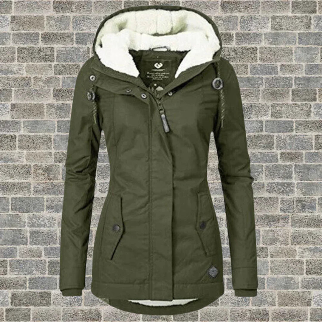 Giacca invernale verde oliva da donna con cappuccio foderato in pelliccia sintetica, chiusura a zip e bottoni, su sfondo di mattoni grigi.