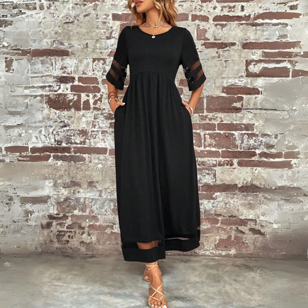 Abito lungo nero elegante da donna con maniche trasparenti, indossato con sandali beige. Sfondo di muro in mattoni. Moda femminile casual chic.