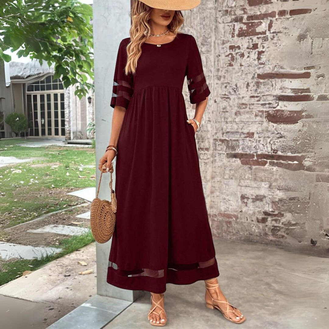 Donna con abito lungo bordeaux, maniche trasparenti, cappello di paglia, borsa rotonda in paglia, sandali beige, sfondo rustico.