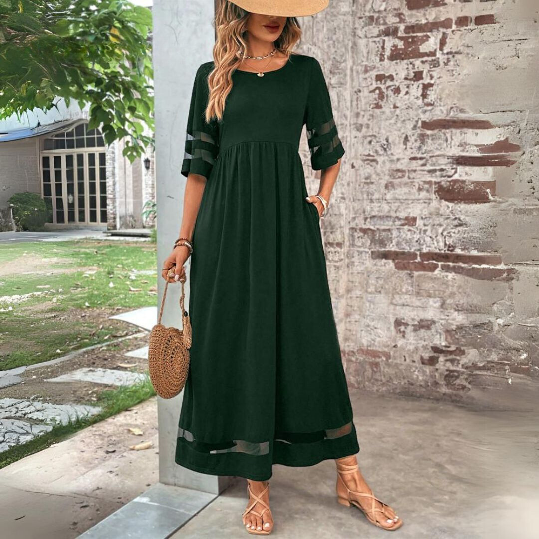 Donna con abito lungo verde scuro, cappello di paglia e borsa rotonda in paglia, posa all'aperto vicino a un muro di mattoni. Moda estiva elegante.