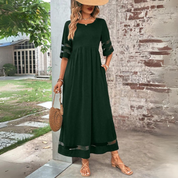 Donna con abito lungo verde scuro, cappello di paglia e borsa rotonda in paglia, posa all'aperto vicino a un muro di mattoni. Moda estiva elegante.