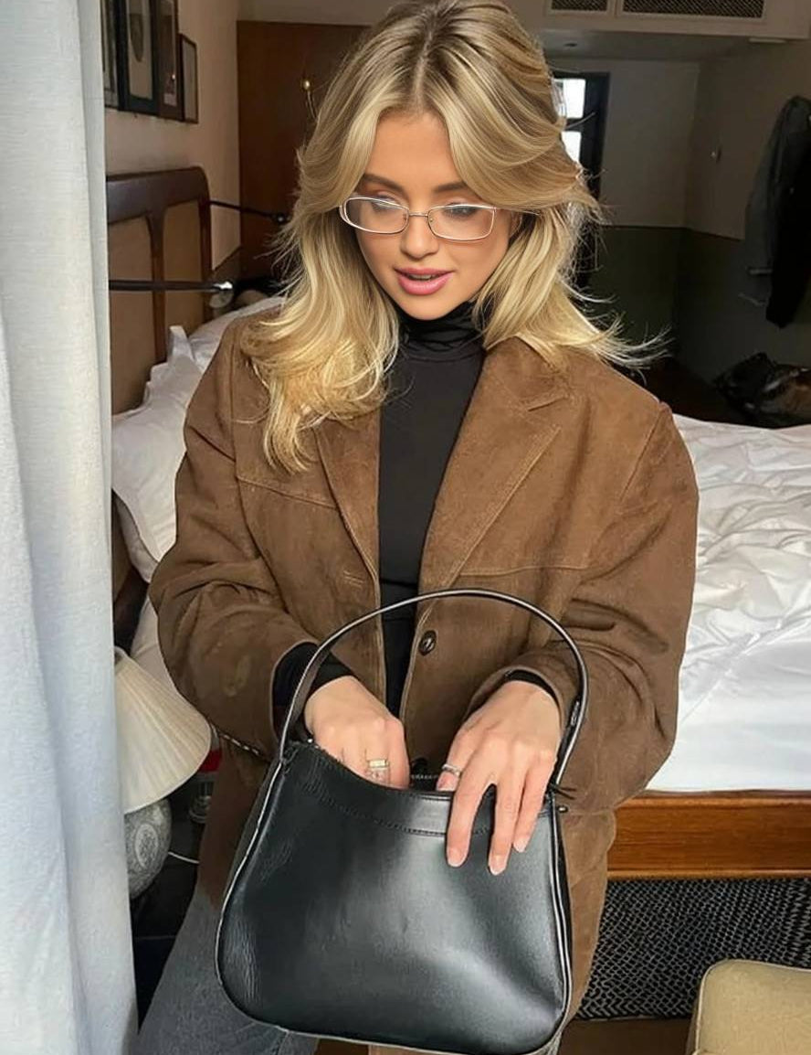Donna con occhiali indossa giacca marrone e tiene borsa nera in camera da letto. Moda autunno, accessori eleganti, stile casual chic.