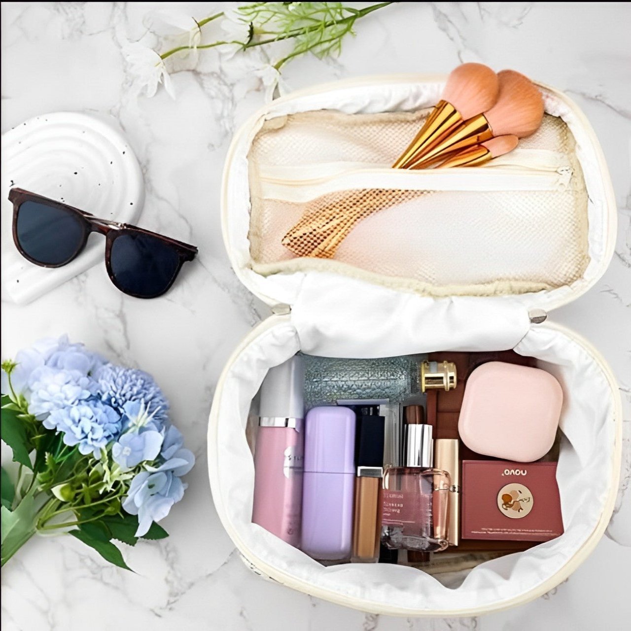 Beauty case aperto su sfondo marmo con trucchi, pennelli trucco, occhiali da sole e fiori blu. Accessori cosmetici eleganti e di tendenza.