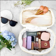 Beauty case aperto su sfondo marmo con trucchi, pennelli trucco, occhiali da sole e fiori blu. Accessori cosmetici eleganti e di tendenza.