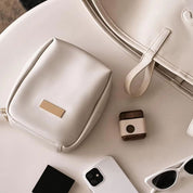 Borsa in pelle bianca su tavolo, con accessori eleganti: custodia, anello in legno, smartphone e occhiali da sole. Stile minimalista e moderno.