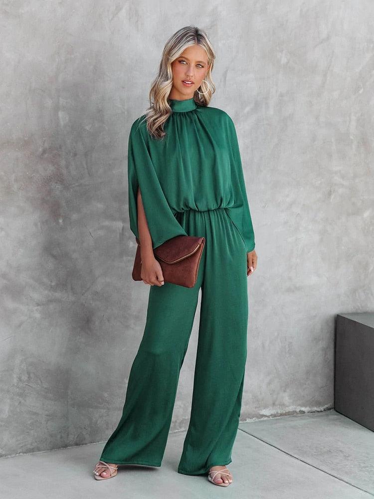 Donna in tuta verde elegante, con pochette marrone e sandali rosa, posa su sfondo grigio. Moda femminile, abbigliamento chic, outfit sofisticato.