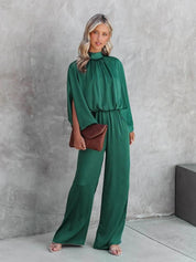 Donna in tuta verde elegante, con pochette marrone e sandali rosa, posa su sfondo grigio. Moda femminile, abbigliamento chic, outfit sofisticato.