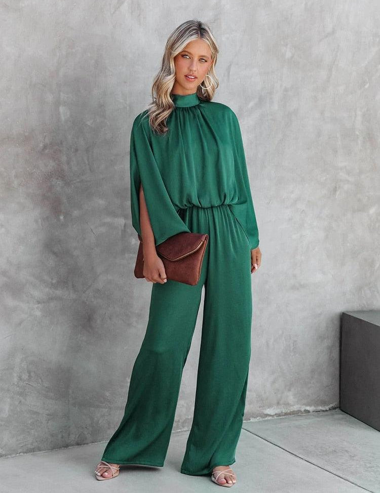 Donna in tuta verde elegante, con pochette marrone e sandali rosa, posa su sfondo grigio. Moda femminile, abbigliamento chic, outfit sofisticato.
