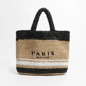 Borsa tote in paglia intrecciata con manici neri, scritta "Paris Milano" e strisce nere e bianche. Accessorio moda estivo elegante.
