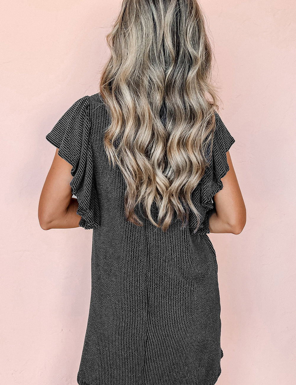 Donna con capelli ondulati biondi indossa un abito nero a coste con maniche corte a volant, sfondo rosa. Moda femminile, abbigliamento estivo elegante.