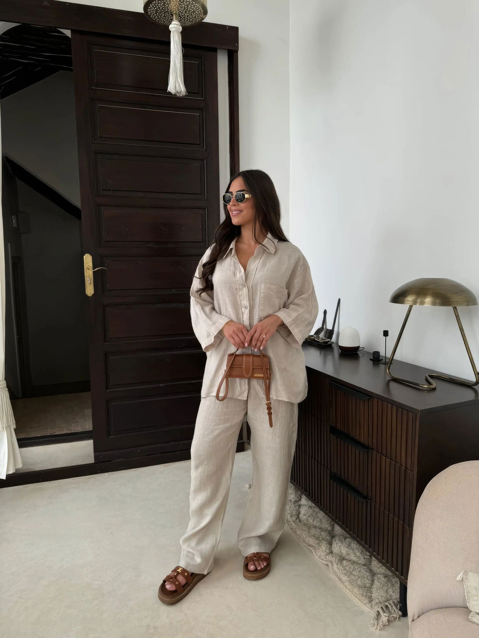 Donna con occhiali da sole indossa camicia e pantaloni beige, sandali marroni, borsa a tracolla. Sfondo interno moderno con porta e lampada.
