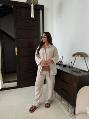 Donna con occhiali da sole indossa camicia e pantaloni beige, sandali marroni, borsa a tracolla. Sfondo interno moderno con porta e lampada.