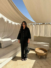 Donna con occhiali da sole indossa abito nero elegante, in terrazza con tende bianche e divani beige. Moda estiva, stile casual chic.