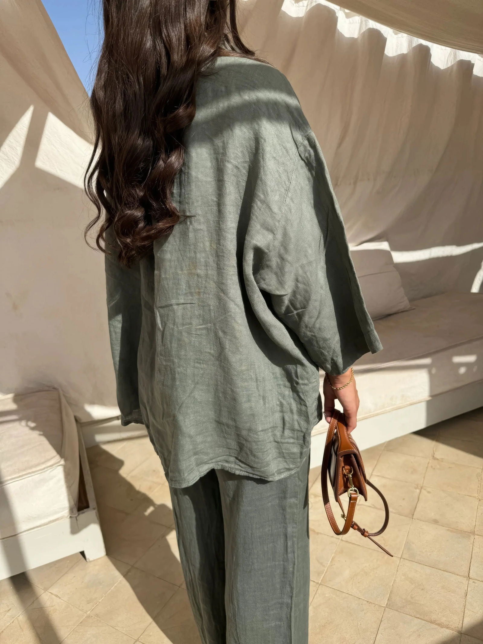 Donna con capelli lunghi indossa un completo in lino verde oliva, giacca e pantaloni, in un ambiente esterno con tende bianche. Moda estiva elegante.