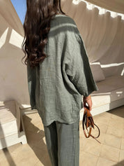 Donna con capelli lunghi indossa un completo in lino verde oliva, giacca e pantaloni, in un ambiente esterno con tende bianche. Moda estiva elegante.