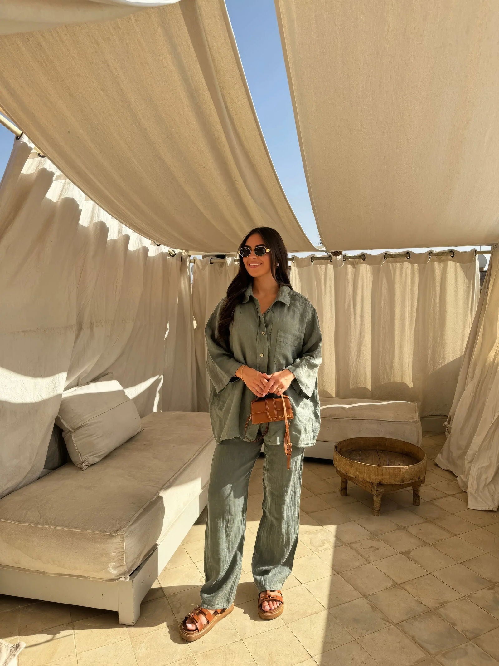 Donna in abbigliamento casual verde oliva, con occhiali da sole e borsa a tracolla marrone, su terrazza con tende beige e divano. Moda estiva elegante.