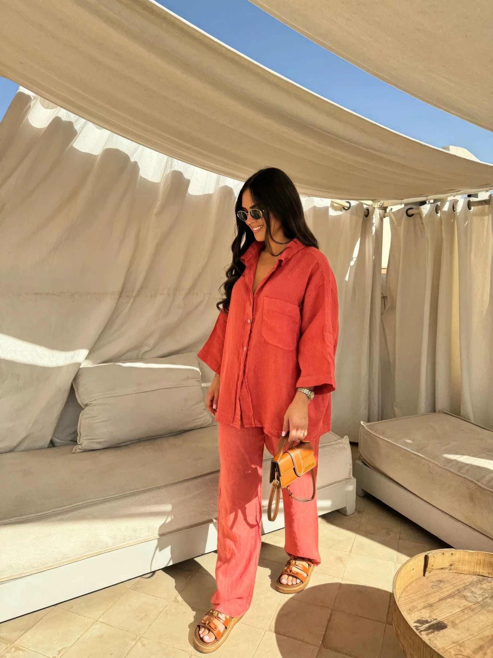 Donna con camicia e pantaloni rossi, borsa a tracolla marrone, sandali, occhiali da sole, in un'area lounge esterna con tende bianche.