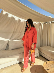 Donna con camicia e pantaloni rossi, borsa a tracolla marrone, sandali, occhiali da sole, in un'area lounge esterna con tende bianche.
