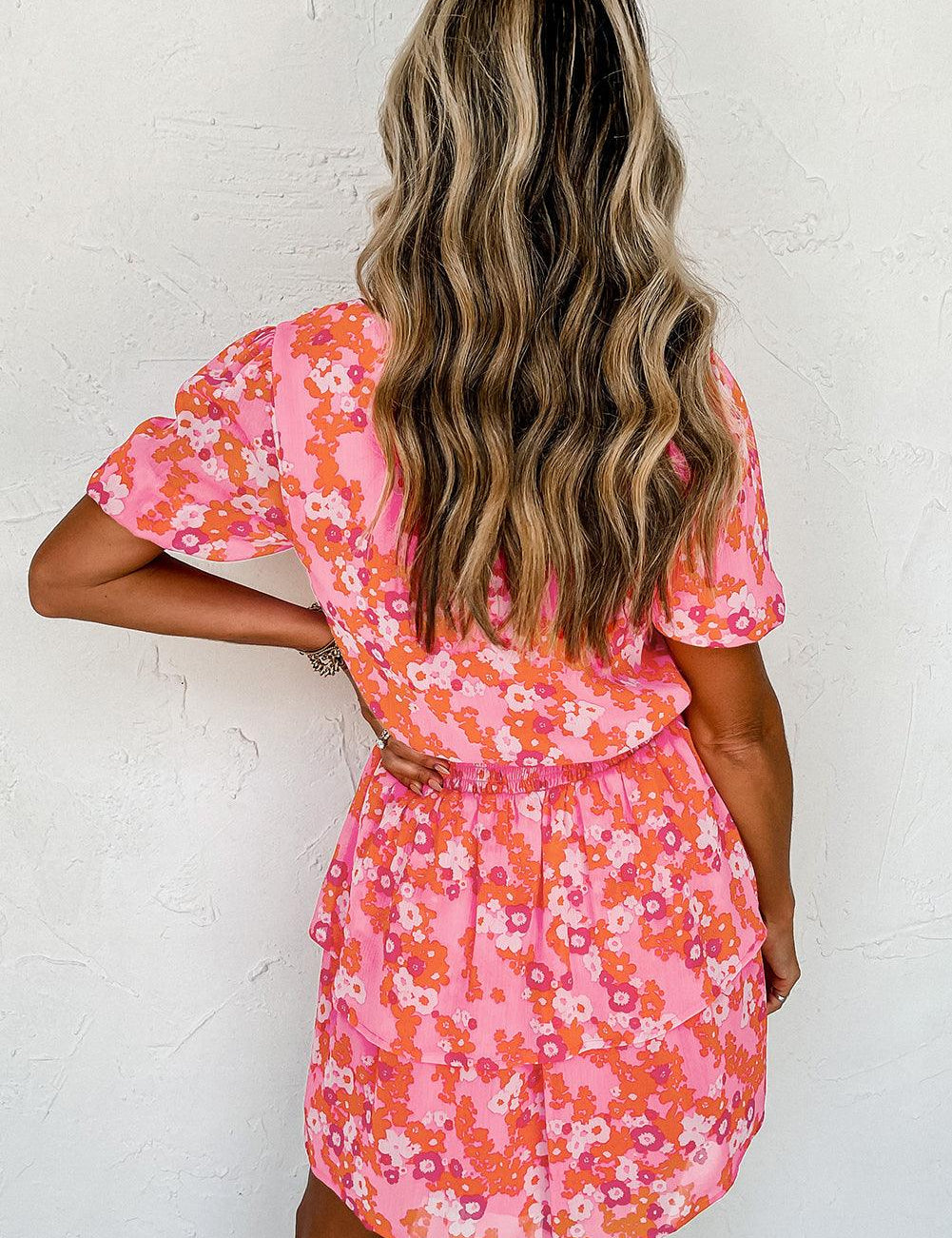 Donna con abito estivo floreale rosa e arancione, capelli ondulati biondi, sfondo bianco. Moda femminile, abbigliamento casual elegante.
